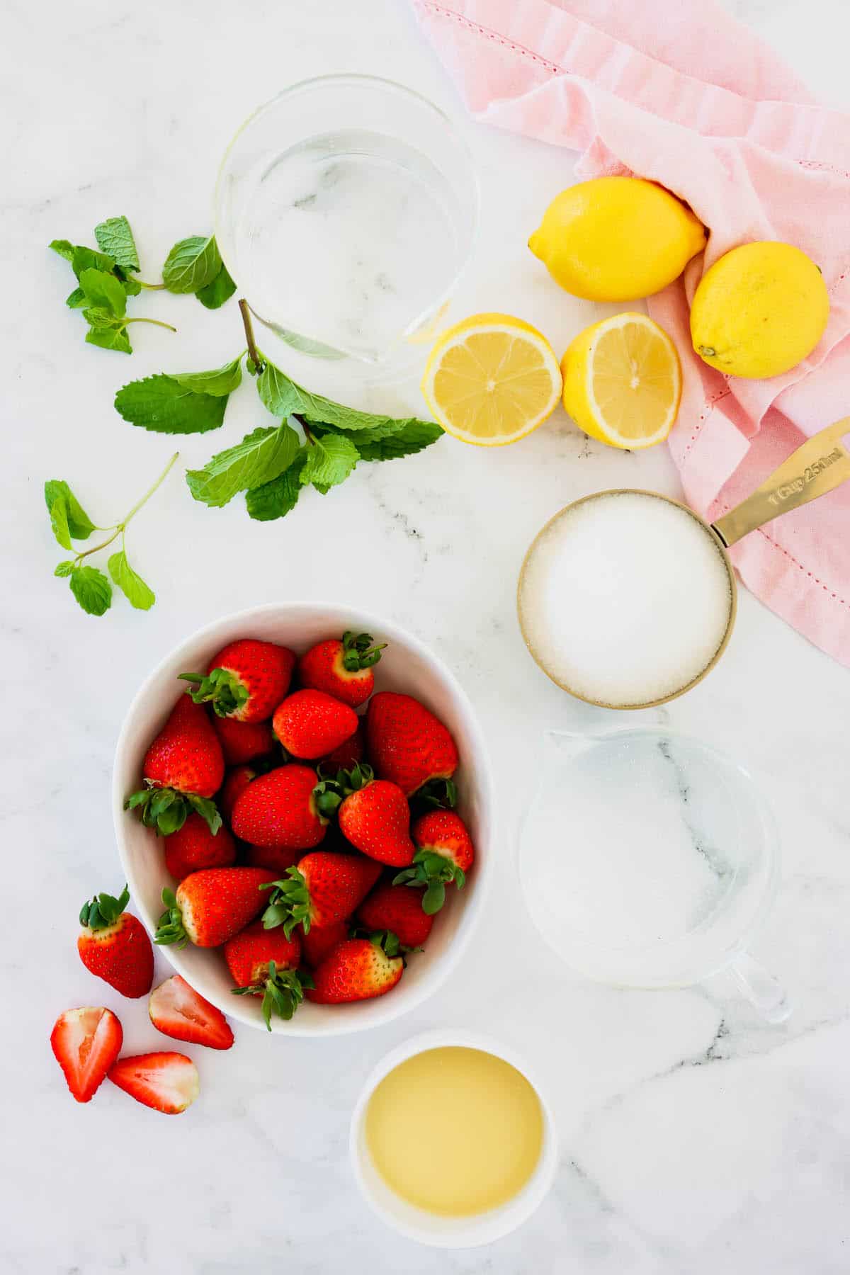 Overhead view of fresh strawberries in a bowl, a measuring cup of sugar and fresh lemons