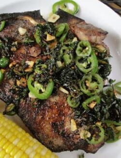 Grilled T-bone steak topped with jalapeno slices and chopped cilantro next to an ear of corn on a white plate