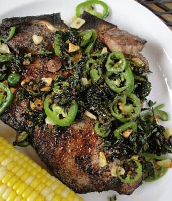 Grilled T-bone steak topped with jalapeno slices and chopped cilantro next to an ear of corn on a white plate