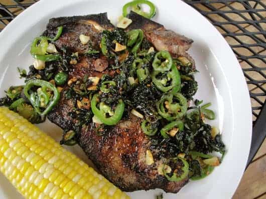 Grilled T-bone steak topped with jalapeno slices and chopped cilantro next to an ear of corn on a white plate