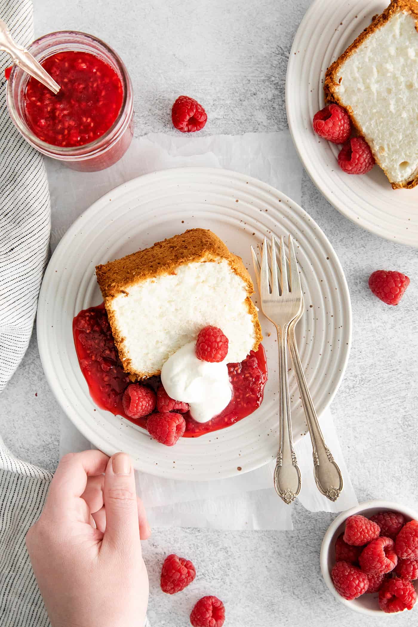 A hand grabbing a slice of angel food cake with whipped cream and raspberry sauce