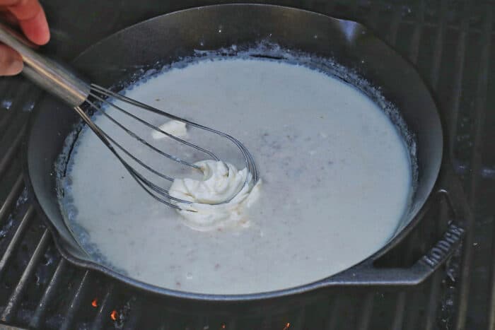 Cream cheese being whisked into a milk and flour mixture in a cast iron skillet