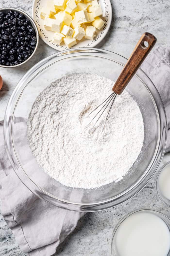 Overhead view of dry ingredients in mixing bowl with whisk, bowl of butter, bowl of dried blueberries, and bowl of buttermilk