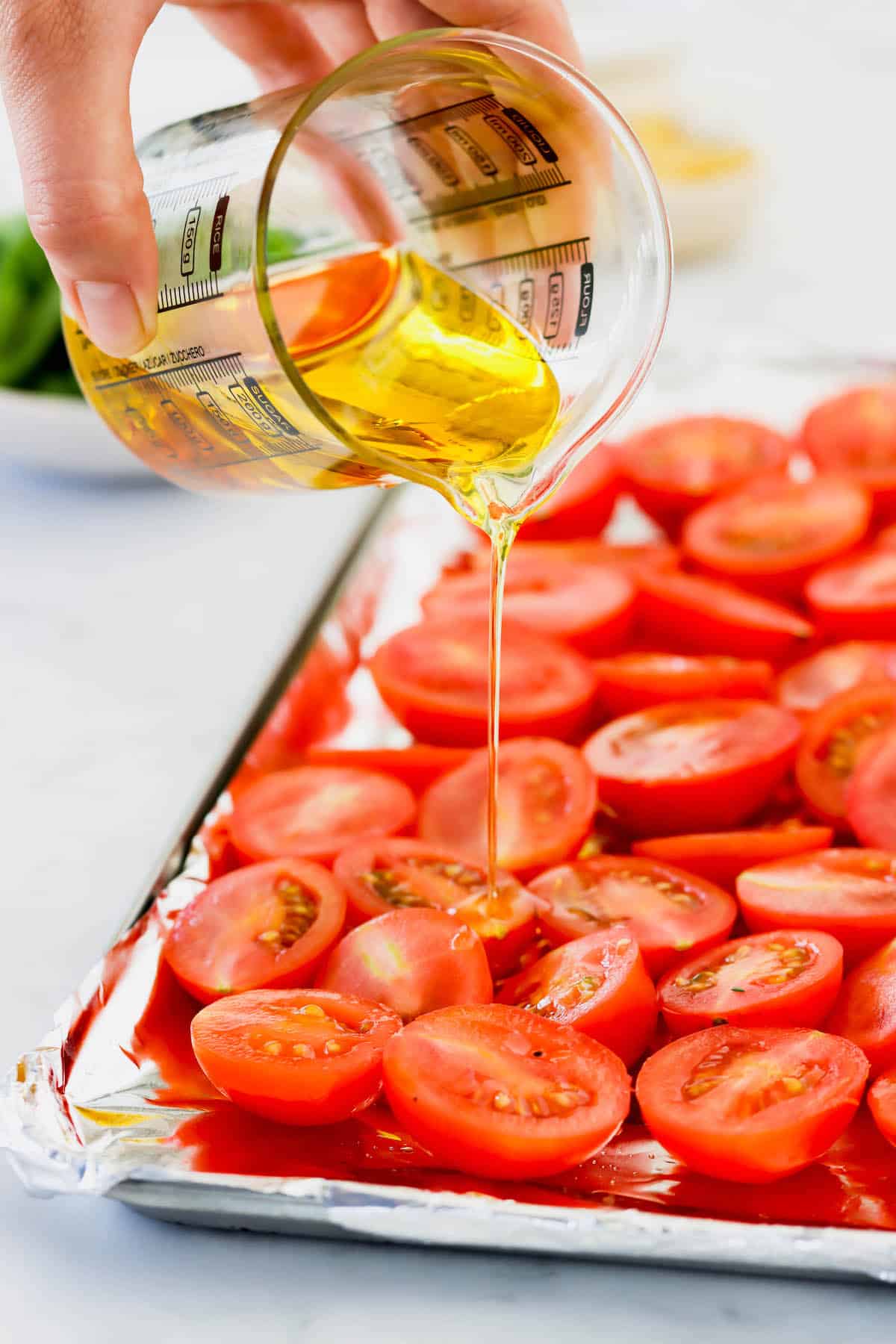 Olive oil being poured over a sheet pan of halved plum tomatoes