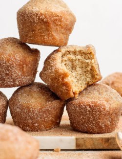 Stack of cinnamon sugar doughnut muffins on wood board, with one muffin bitten into to show moist interior