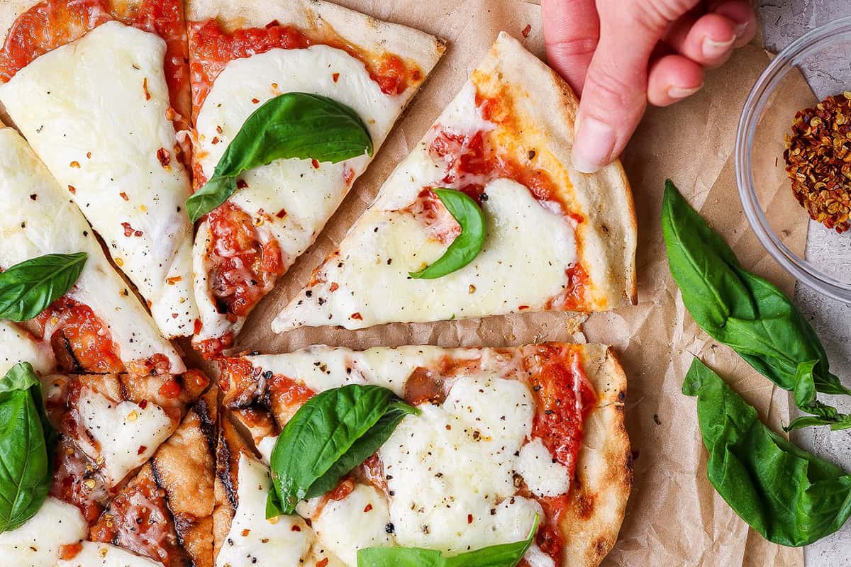 This Grilled Pizza recipe is tested and perfected, with a homemade dough that doesn't fall through the grates. Smoky, crispy, and delicious! Hand pulling slice of grilled Margherita pizza from pie.