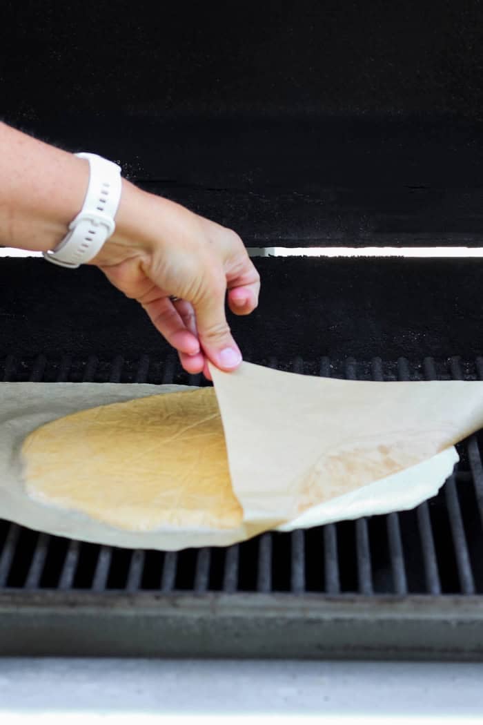 This Grilled Pizza recipe is tested and perfected, with a homemade dough that doesn't fall through the grates. Smoky, crispy, and delicious! Pulling parchment paper off of dough for grilled pizza crust.
