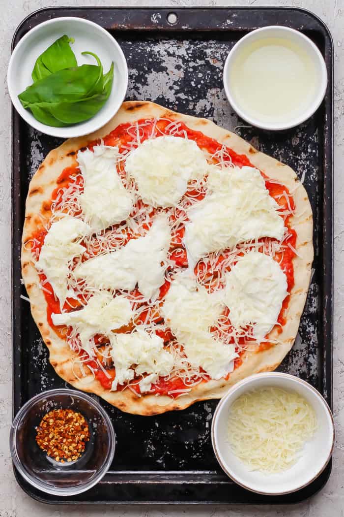 This Grilled Pizza recipe is tested and perfected, with a homemade dough that doesn't fall through the grates. Smoky, crispy, and delicious! Overhead view of grilled margherita pizza on sheet pan with bowls of toppings.