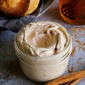 A jar of whipped cinnamon honey butter with a basket of popovers and a jar of honey in the background.