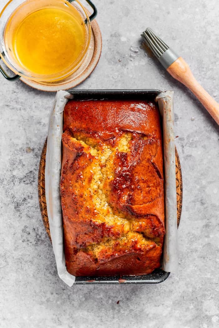 Loaf of orange poppy seed bread in pan with bowl of glaze and brush.