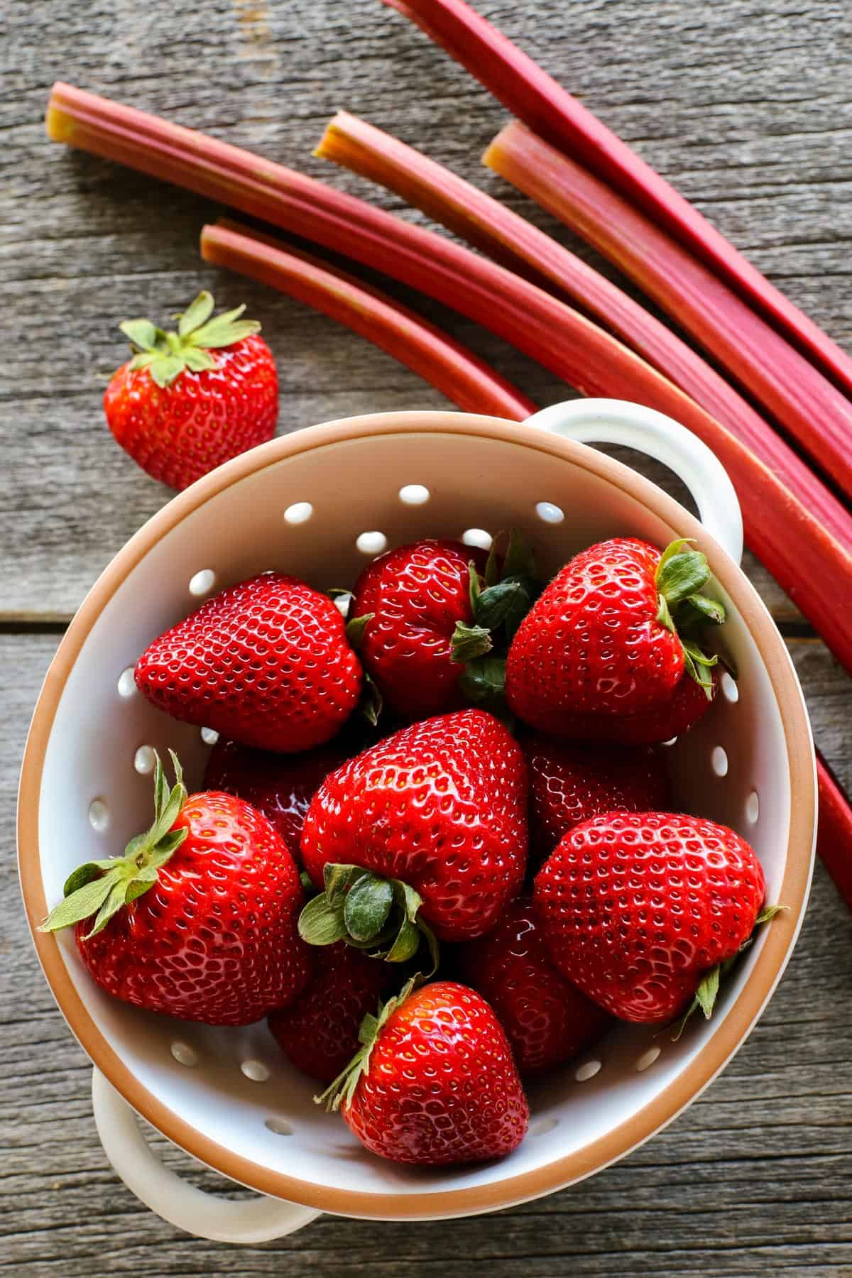 Strawberries are placed in in a colander alongside stalks of rhubarb.