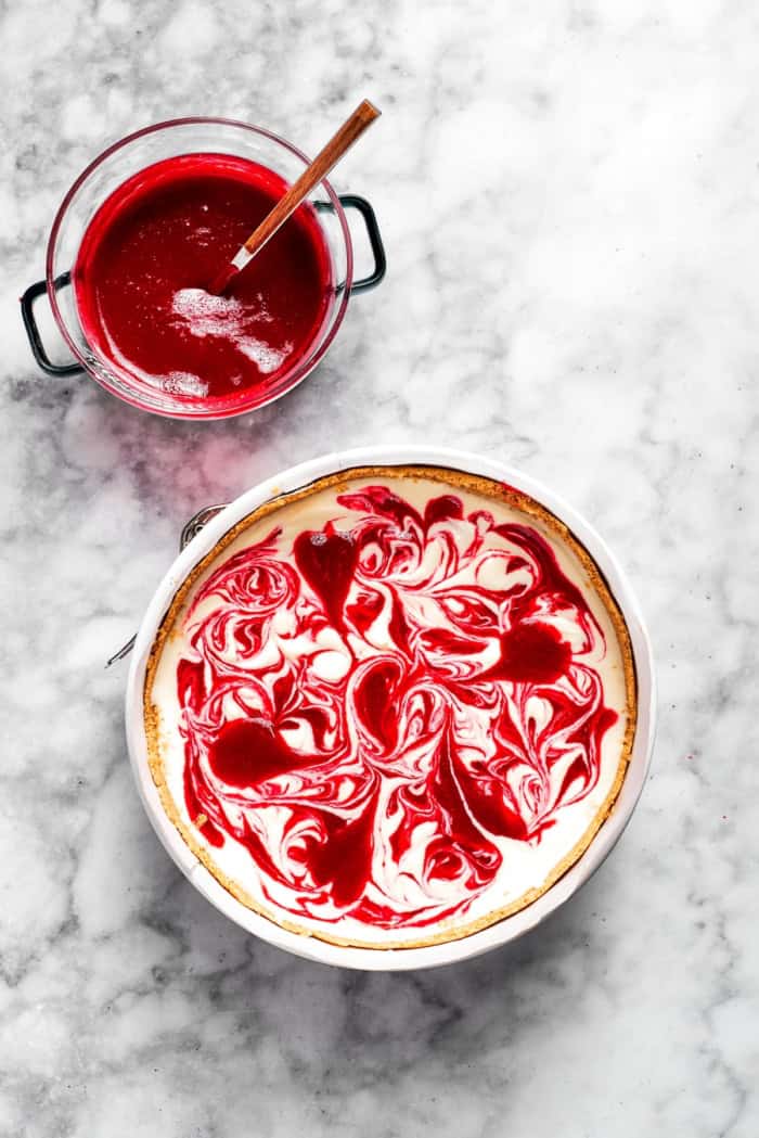 This rich raspberry cheesecake is sure to make your all-time-favorites list. It's rich, creamy, and elegant, but surprisingly easy to make! Overhead view of raspberry swirl cheesecake before baking with bowl of raspberry sauce