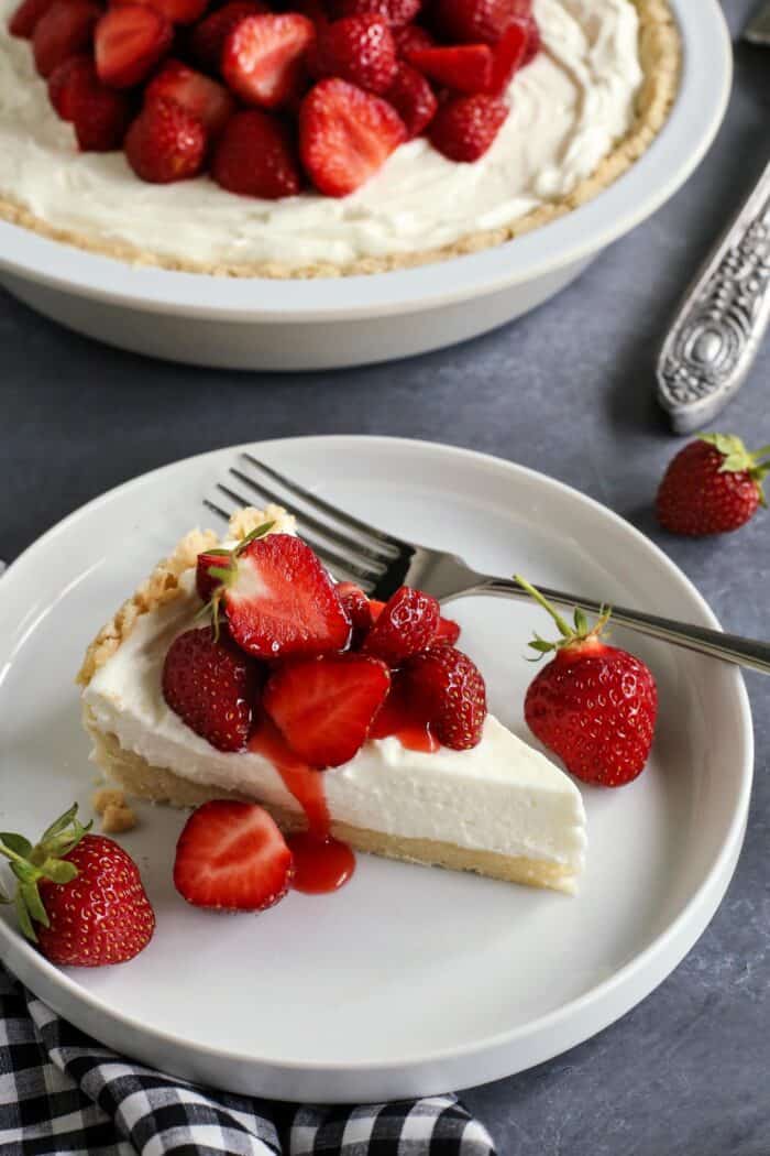 a slice of cream cheese pie topped with fresh strawberries, plus a whole pie in the background