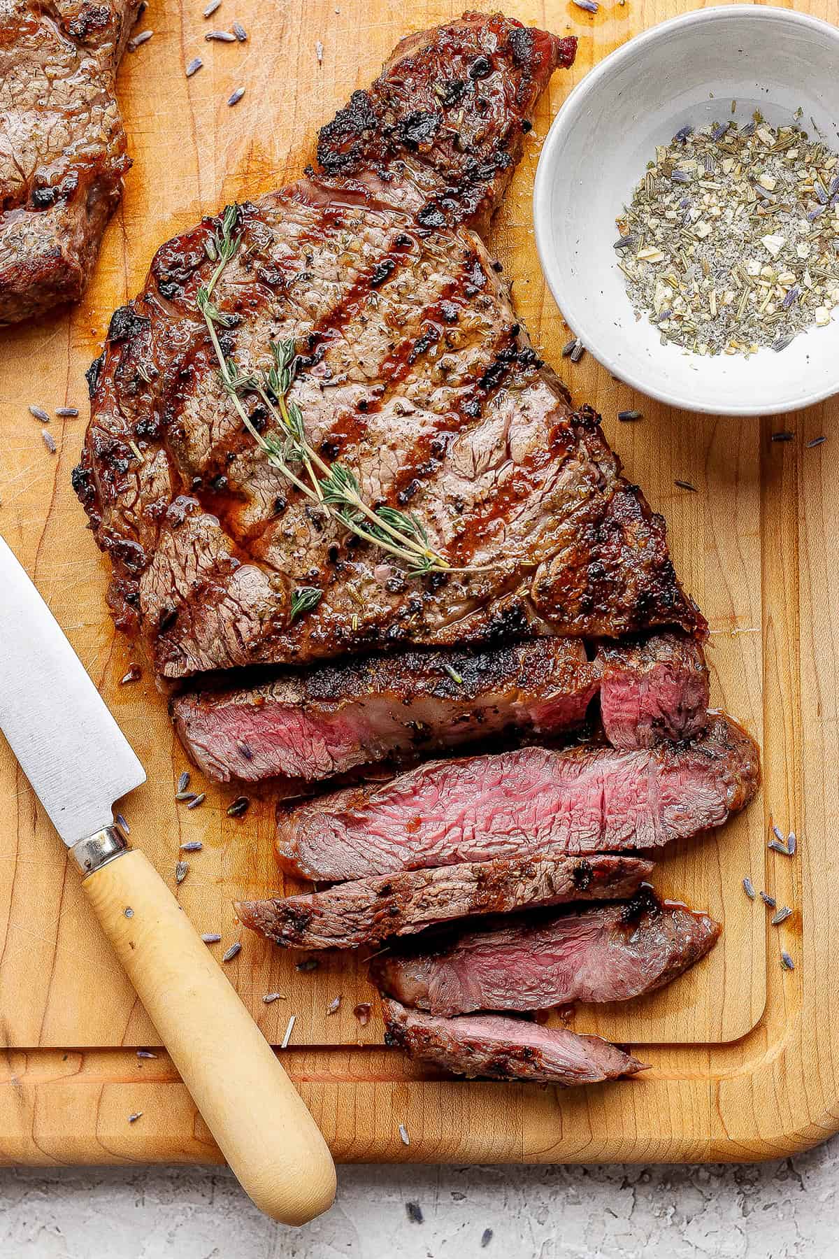Sliced grilled ribeye steak on cutting board with knife and bowl of steak seasoning.