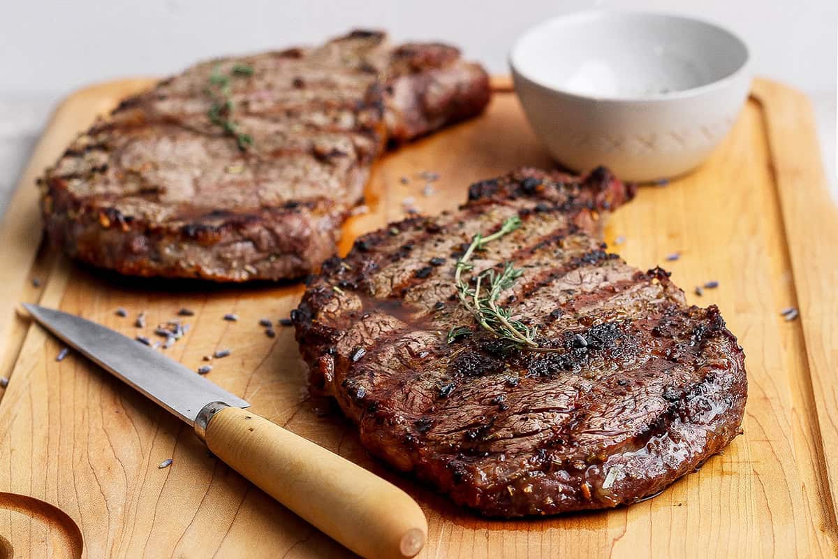 Two grilled ribeye steaks on cutting board with knife.