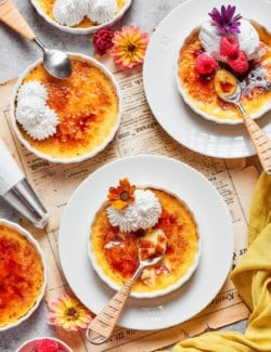4 ramekins of Almond Vanilla Crème Brûlée topped with whipped cream, berries, and flowers.