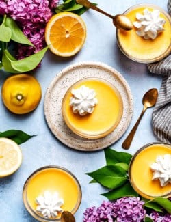 Overhead view of 4 lemon custard cups with whipped cream, surrounded by fresh lemons and lilacs