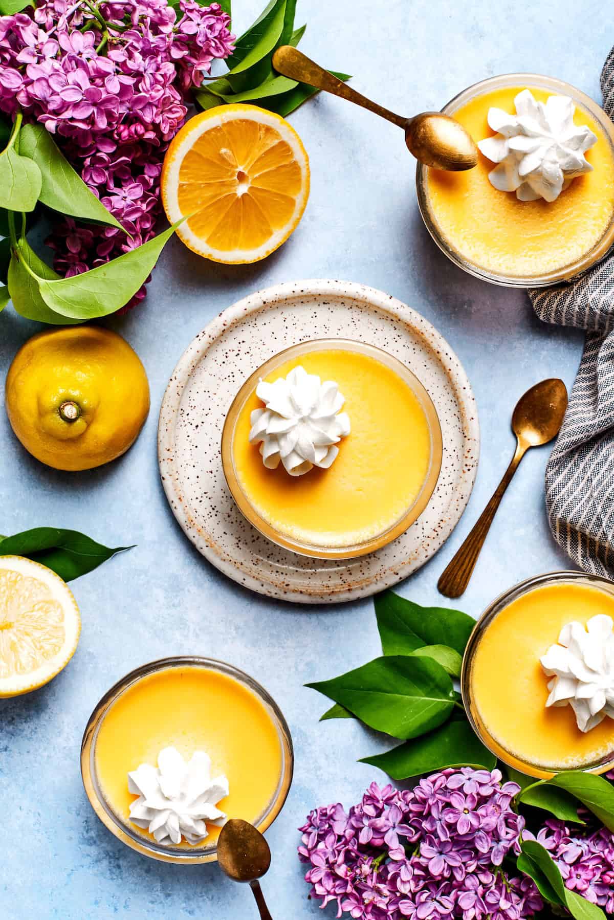 These mini lemon custard cups have a delicate lemon flavor and a smooth, creamy texture that makes them irresistible. They're a delight! Overhead view of 4 lemon custard cups with whipped cream, surrounded by fresh lemons and lilacs