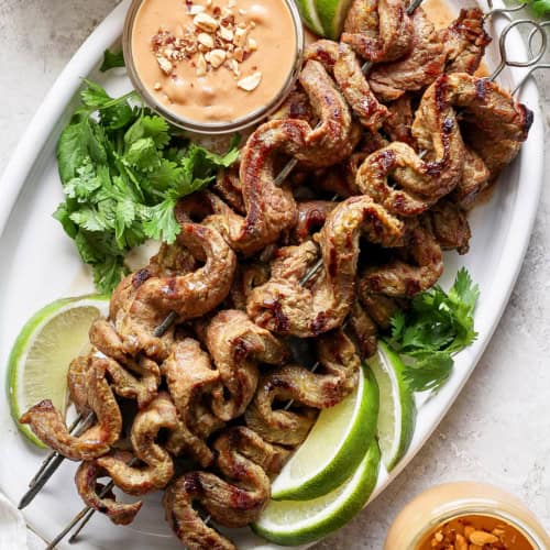 Overhead view of coconut beef satay on platter with bowl of peanut sauce, cilantro, and lime wedges