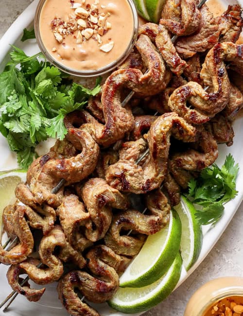 Overhead view of coconut beef satay on platter with bowl of peanut sauce, cilantro, and lime wedges