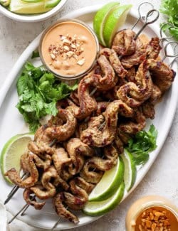 Overhead view of coconut beef satay on platter with bowl of peanut sauce, cilantro, and lime wedges