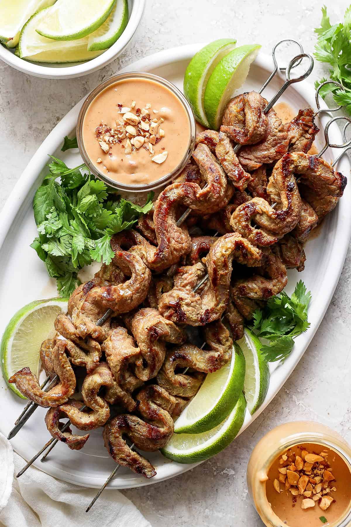 Tender beef is marinated in coconut milk, threaded onto skewers, and grilled to perfection in this coconut beef satay recipe. So flavorful! Overhead view of coconut beef satay on platter with bowl of peanut sauce, cilantro, and lime wedges