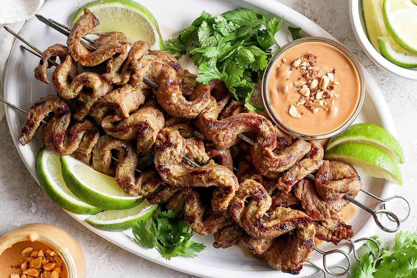 Overhead view of coconut beef satay on platter with bowl of peanut sauce, cilantro, and lime wedges