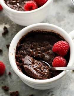 a spoon dipping into a white mug of chocolate cake with fresh raspberries
