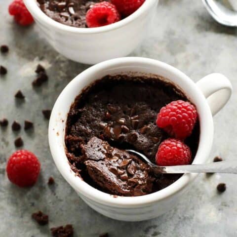 a spoon dipping into a white mug of chocolate cake with fresh raspberries