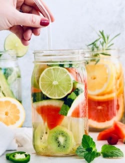Hand holding glass straw in jar of spa water with kiwi, lime, and watermelon