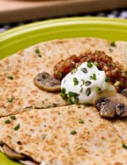 A round quesadilla on a plate topped with sliced sauteed mushrooms, sour cream, salsa, scallions and a lime wedge