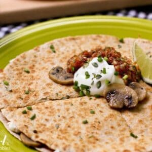 A round quesadilla on a plate topped with sliced sauteed mushrooms, sour cream, salsa, scallions and a lime wedge