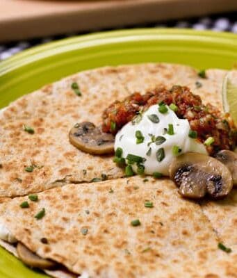 A round quesadilla on a plate topped with sliced sauteed mushrooms, sour cream, salsa, scallions and a lime wedge