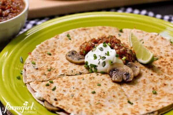 A round quesadilla on a plate topped with sliced sauteed mushrooms, sour cream, salsa, scallions and a lime wedge