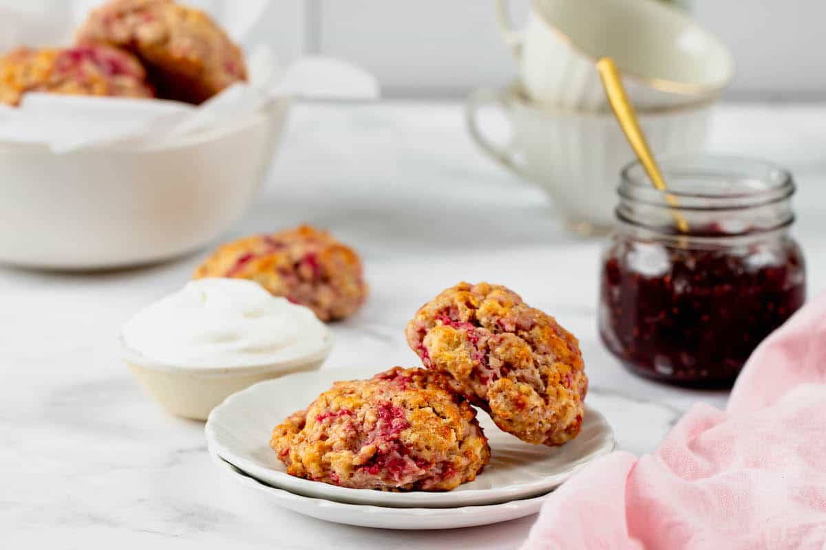 Two raspberry drop scones on a plate with jars or cream cheese and jelly behind them. 