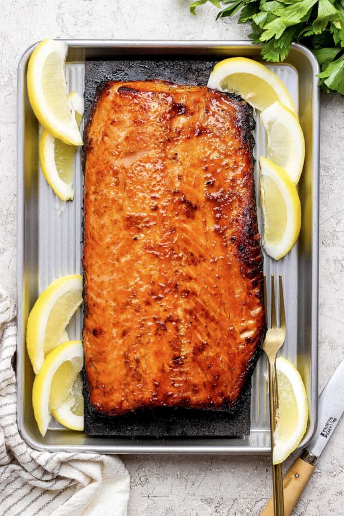 Overhead view of sweet and smoky grilled salmon set on cedar plank inside sheet pan with lemon wedges
