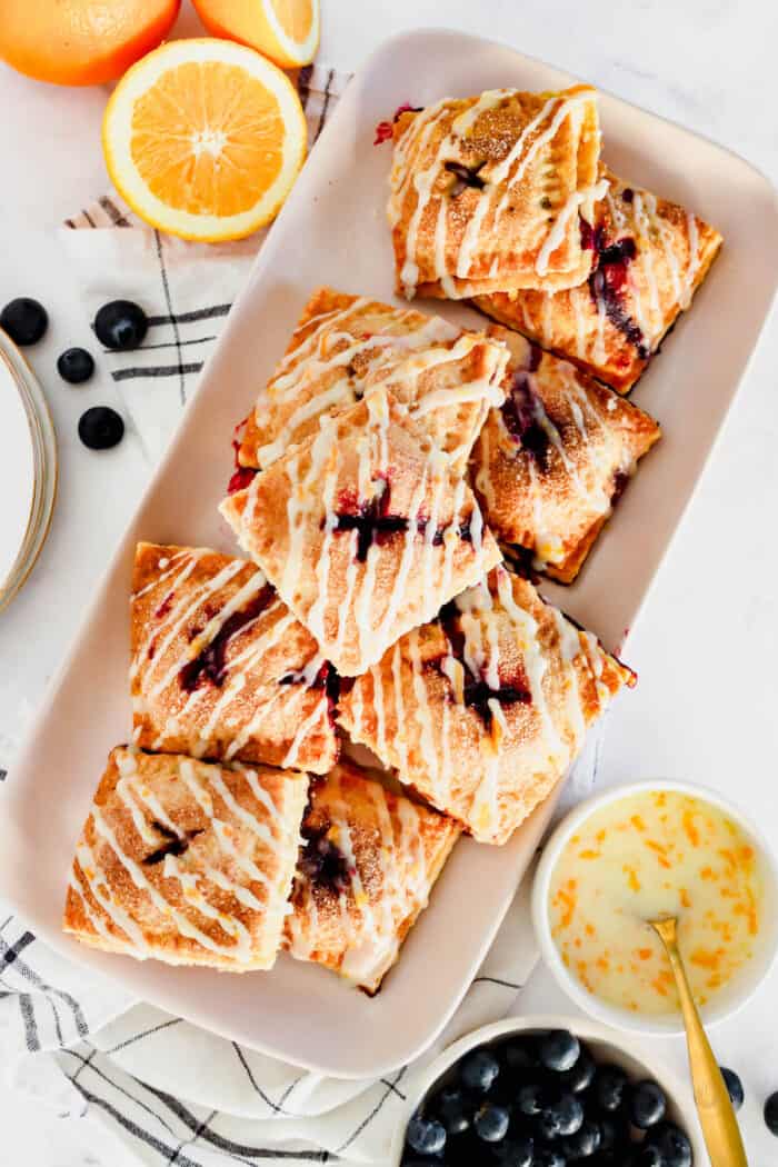 Overhead view of a platter of blueberry hand pies drizzled with orange glaze, next to small bowls with pie ingredients.