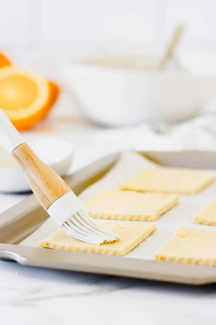 A basting brush brushing egg wash over squares of cut out pie dough on a lined baking sheet.