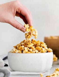 A hand grabbing a piece of caramel popcorn from a white bowl