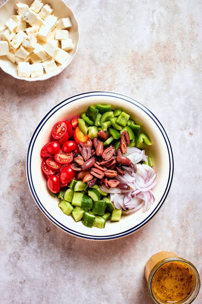 This Greek Chopped Salad is loaded with fresh cucumbers, tomatoes, bell peppers, and herbs and drizzled with a bright, flavorful dressing. Overhead view of bowl of feta, bowl of chopped vegetables, and jar of Greek dressing