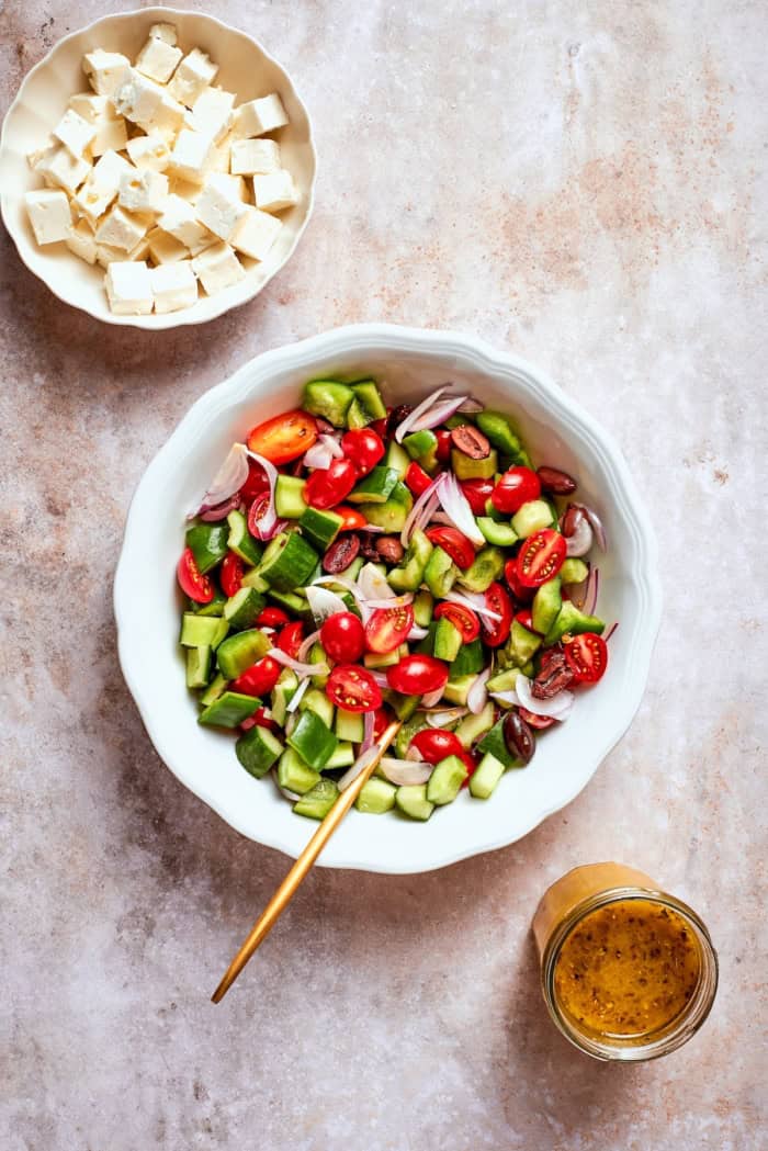 This Greek Chopped Salad is loaded with fresh cucumbers, tomatoes, bell peppers, and herbs and drizzled with a bright, flavorful dressing. Overhead view of bowl of feta, bowl of chopped vegetables, and jar of Greek dressing