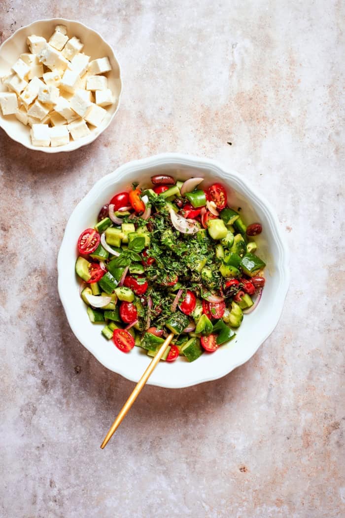 This Greek Chopped Salad is loaded with fresh cucumbers, tomatoes, bell peppers, and herbs and drizzled with a bright, flavorful dressing. Overhead view of fresh herbs added to bowl of Greek salad and bowl of feta