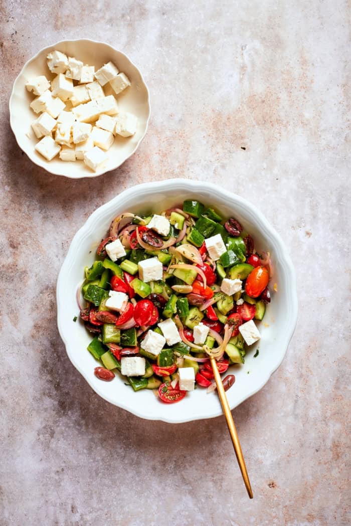 This Greek Chopped Salad is loaded with fresh cucumbers, tomatoes, bell peppers, and herbs and drizzled with a bright, flavorful dressing. Overhead view of Greek chopped salad in bowl set next to bowl of feta