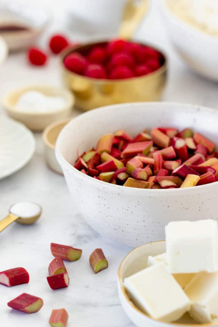 A bowl of chopped rhubarb with cubed butter and raspberries in the background