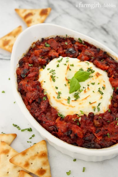 An oval baking dish of baked goat cheese surrounded by sun-dried tomato dip