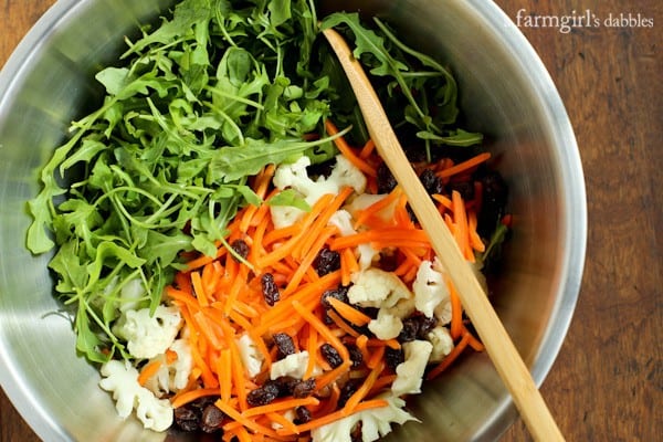 Arugula, carrots, cauliflower and raisins in a mixing bowl with a wooden spoon