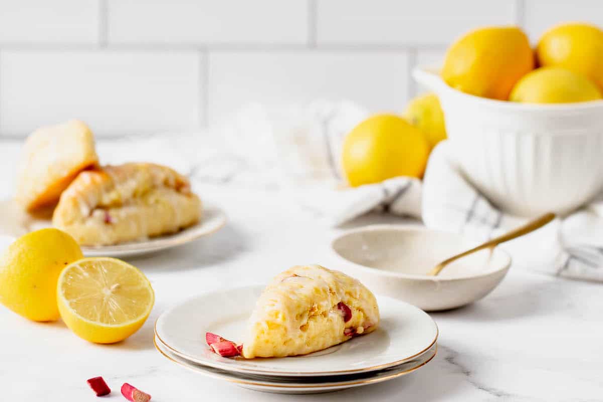 Two lemon scones on white plates, with a white bowl of lemons in the background