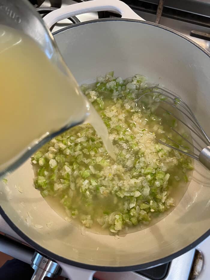 Seafood stock being added to a soup pan with scallions and garlic