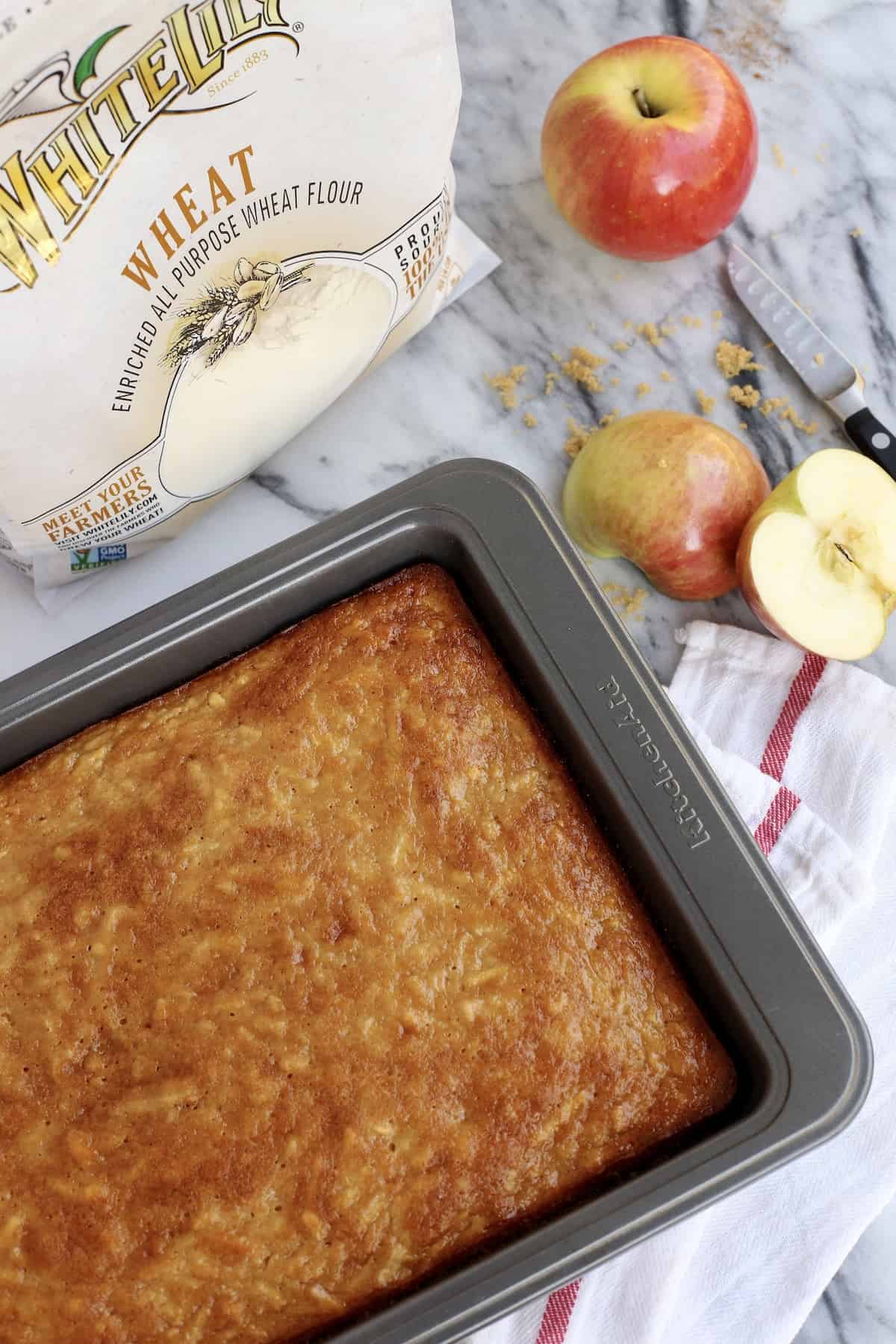 Unfrosted apple cake in a pan next to a bag of flour and apples