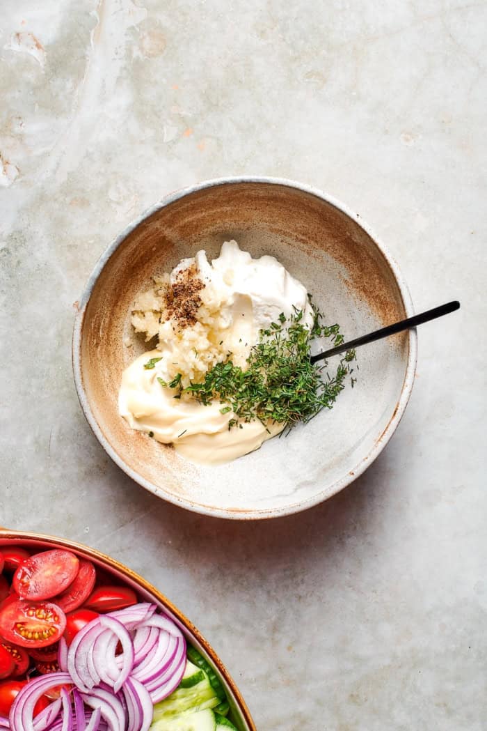 With fresh vegetables and a creamy herbed dressing, this cucumber and tomato salad is the perfect goes-with-anything side dish for summer. Ingredients for creamy dressing in bowl with spoon.